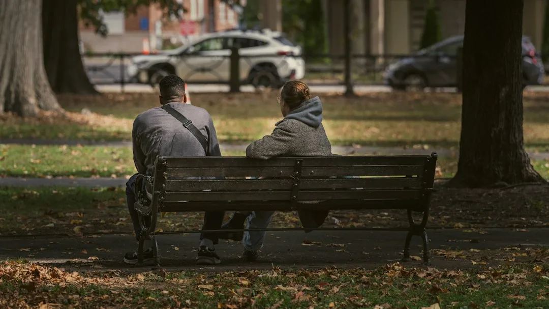 Two people sitting side by side on a park bench, suggesting quiet companionship and listening.
